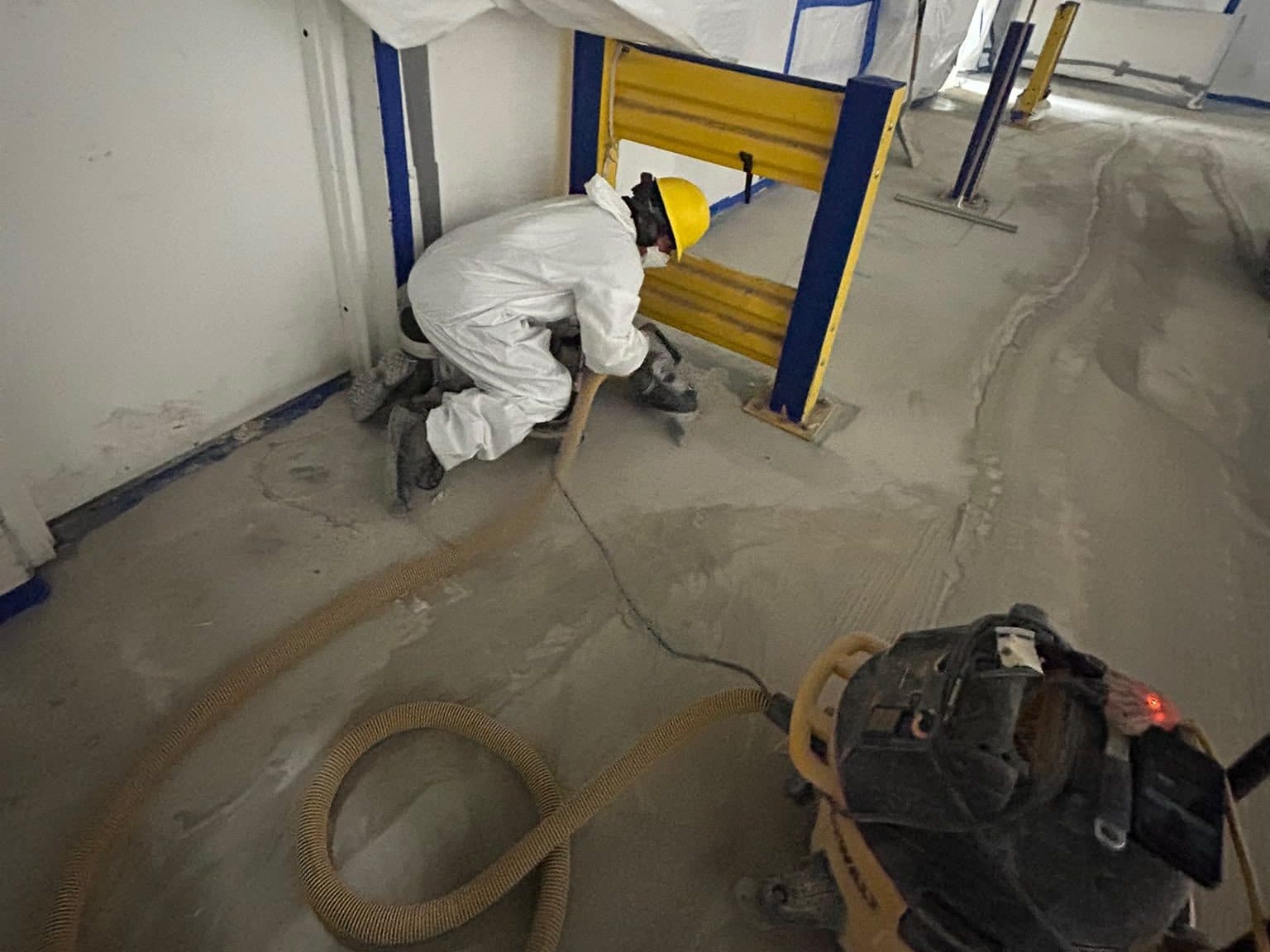 Crew member in disposable coveralls, hard hat and respirator edge-grinding a concrete floor beside a guardrail, with a dust shroud and vacuum hose inside a poly enclosure